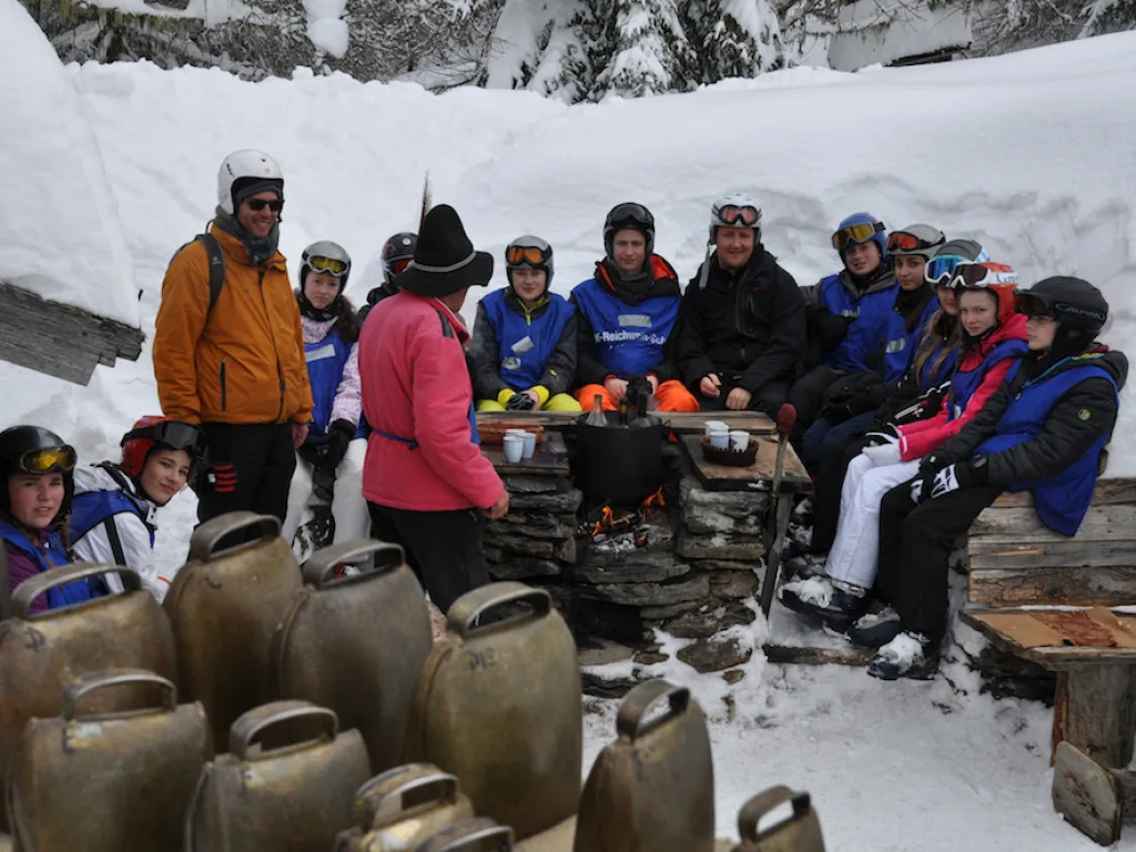 Gruppe sitzt an einem Holztisch vor der Hütte, davor Kuhglocken.